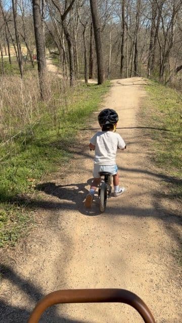 A young child in a helmet riding a balance bike down a wooded trail in Northwest Arkansas